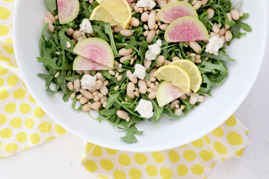 Yellow napkin with white bowl on top filled with arugula salad. Topped with pine nuts, goat cheese and sliced radishes.