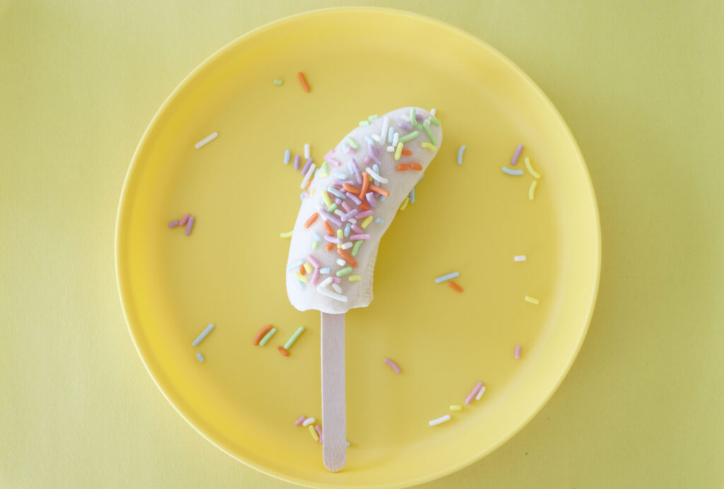 Banana half covered in yogurt with rainbow sprinkles on top on a popsicle stick on a yellow plate on top of yellow paper