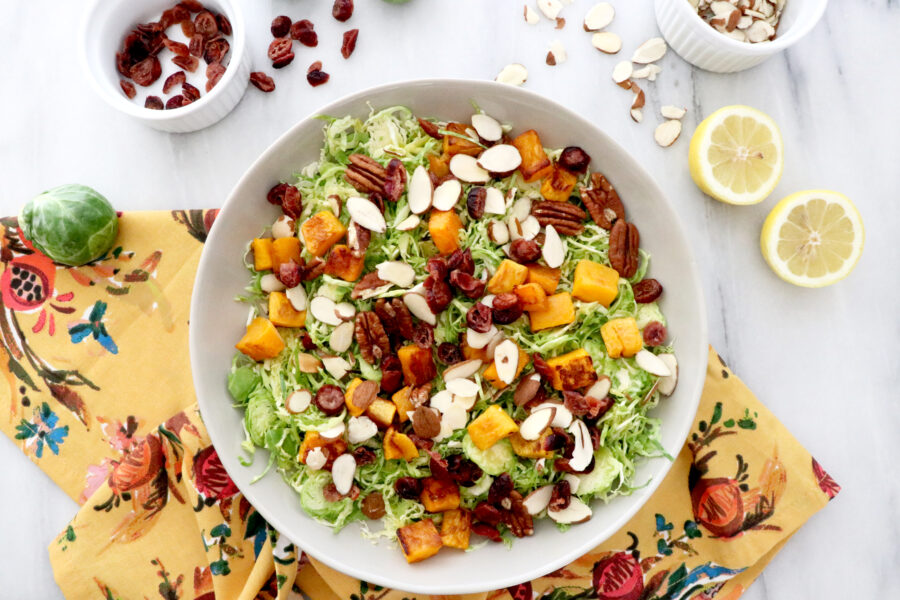Large white bowl filled with Brussels sprouts butternut squash salad with cranberries all on a colorful napkin. Lemons and cranberries on the side.