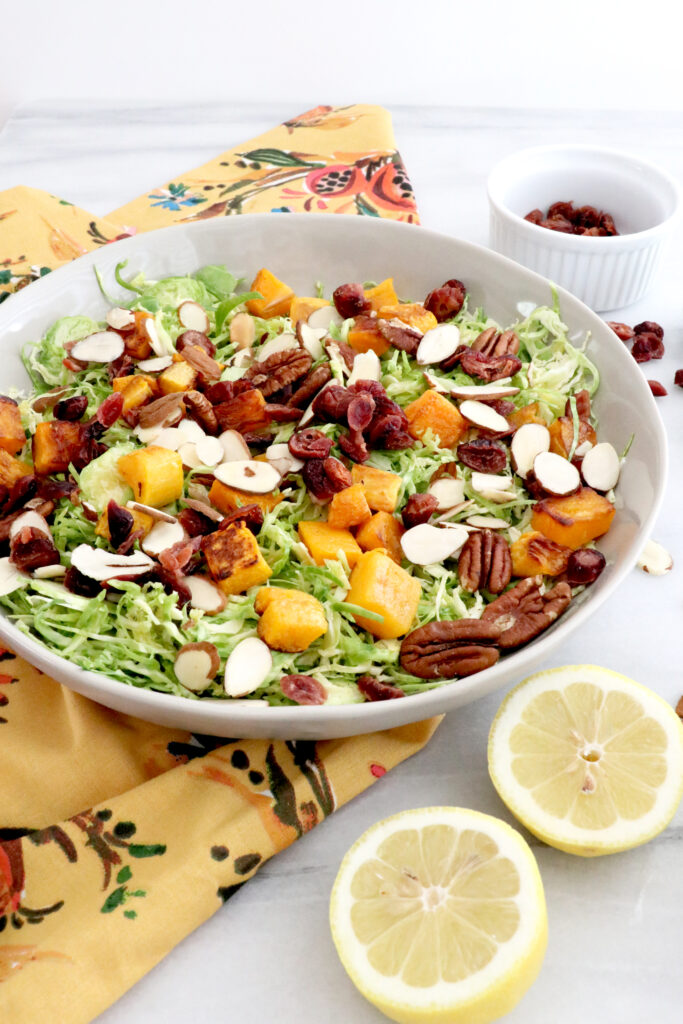 Large white bowl filled with Brussels sprouts butternut squash salad with cranberries all on a colorful napkin. Lemons and cranberries on the side.