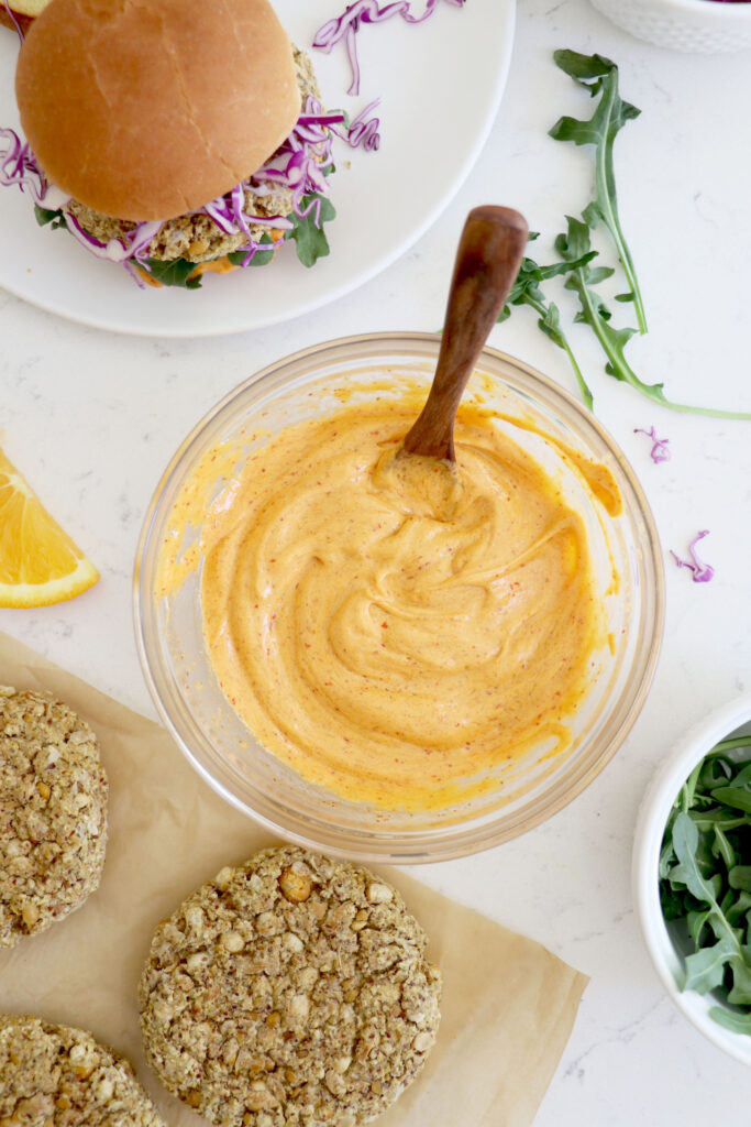 Bowl of buffalo sauce, chickpea burgers in left bottom corner and full burger with bun in top left corner.