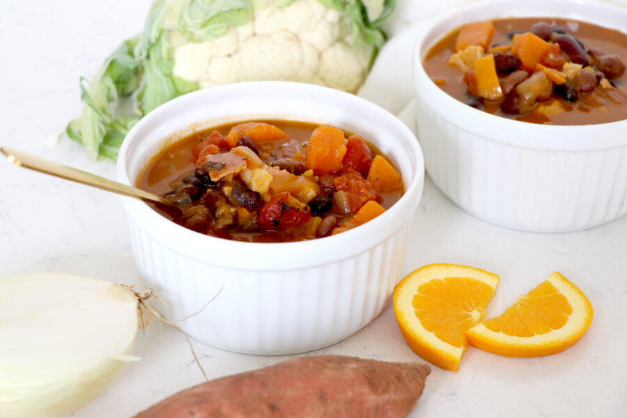 Two white bowls of sweet potato chili surrounded by orange slices, cauliflower and sweet potato.