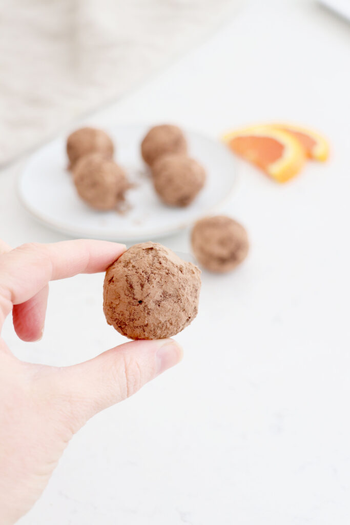 Hand holding truffle with a plate in the background of Chocolate truffles and orange slices.