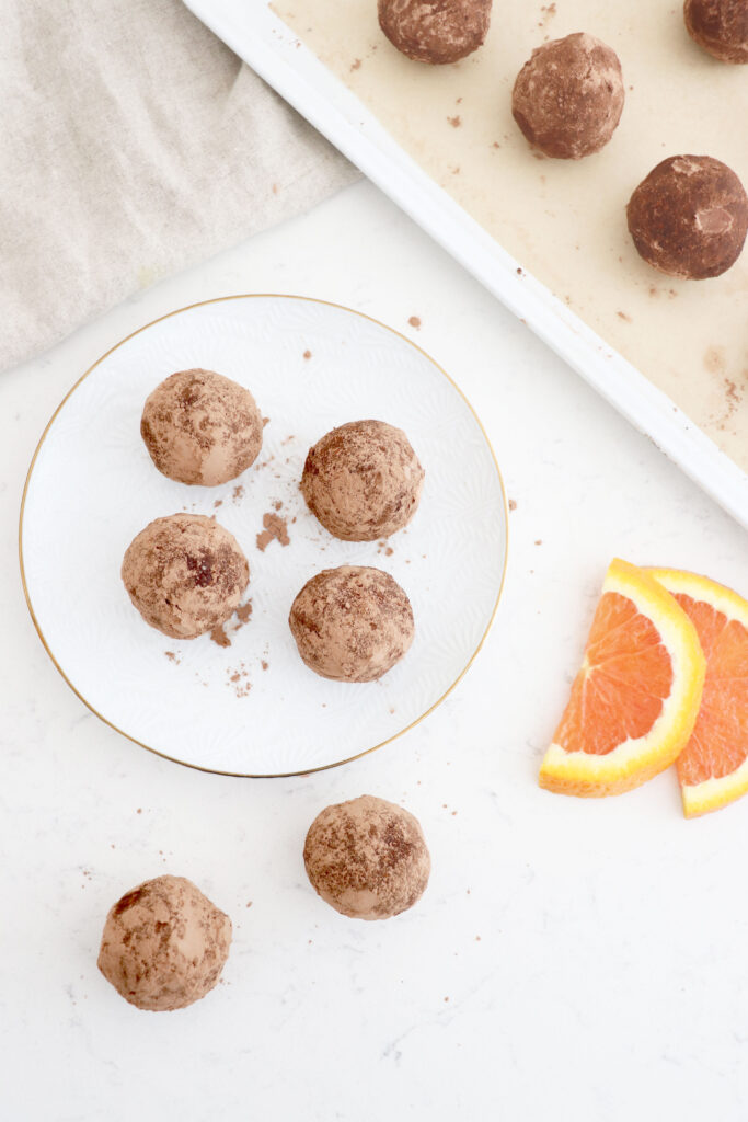 White plate of chocolate truffles with some on both sides. More truffles on baking sheet next to plate.