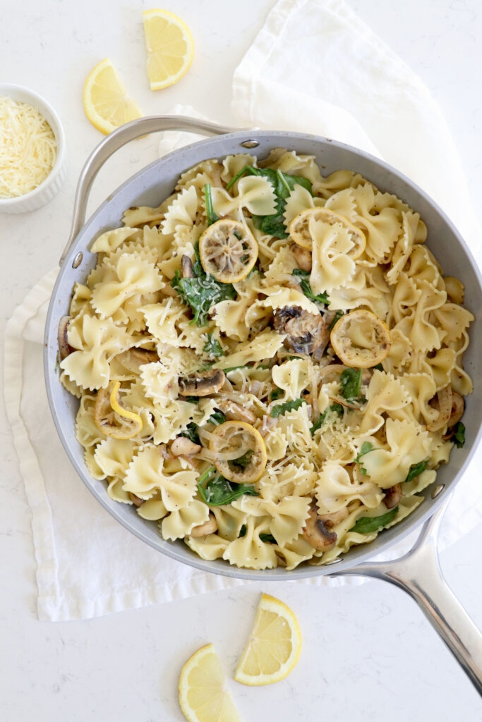 Saucepan with bowtie pasta, lemon slices, spinach and mushrooms.
