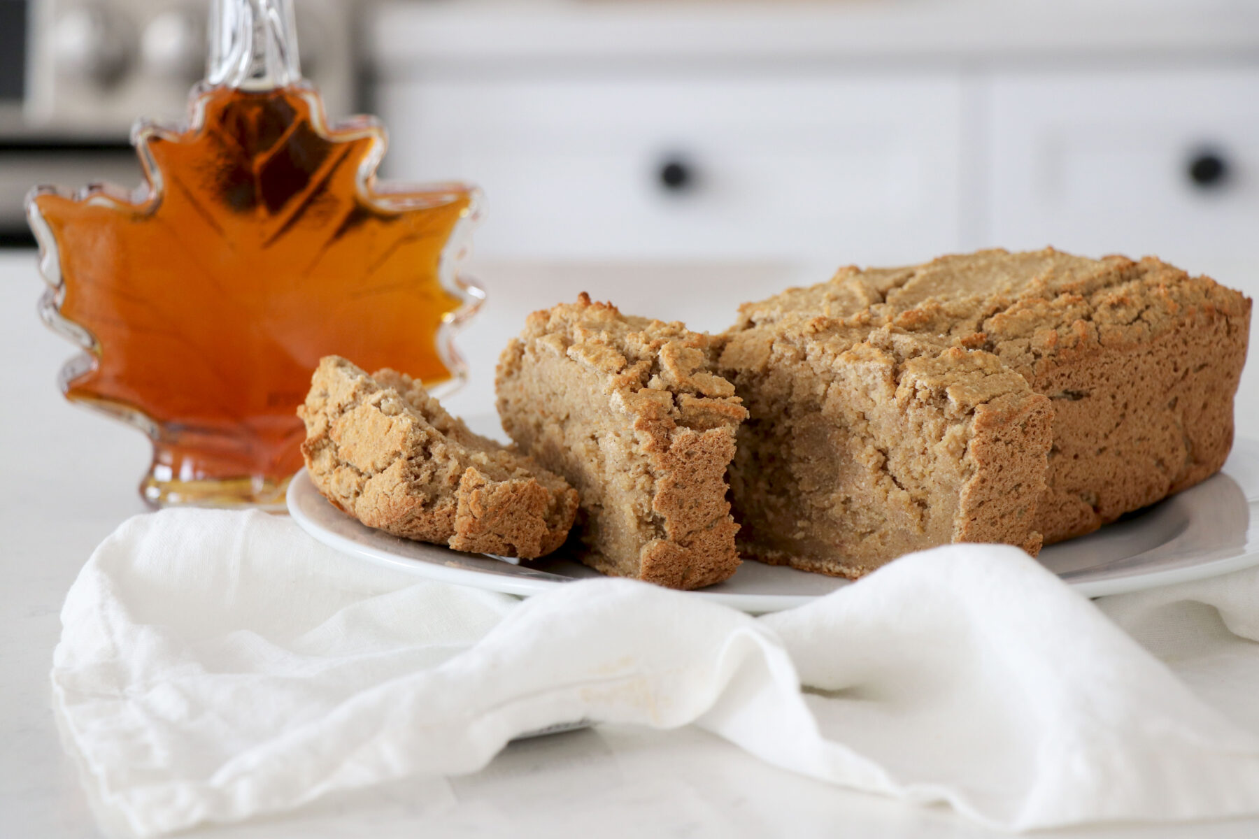 Gingerbread loaf on white plate with white napkin underneath. Maple syrup bottle on the left.