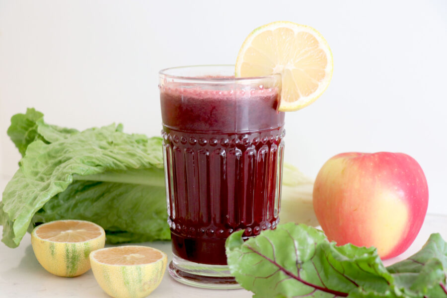 Glass with beet juice surrounded by lettuce, apple and sliced lemon.