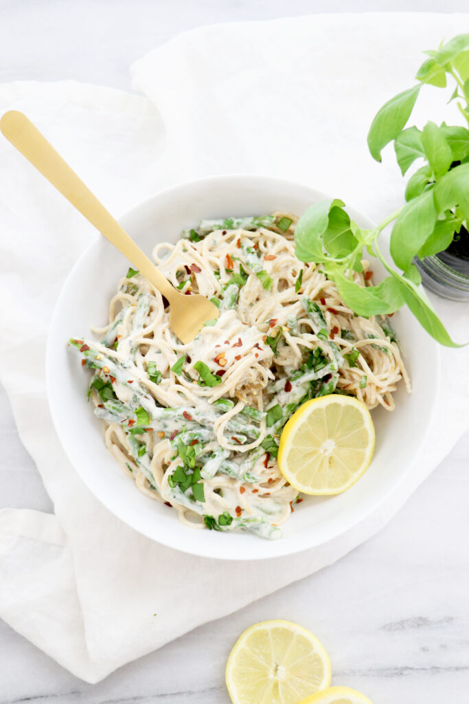 White bowl filled with pasta, cashew cream sauce and basil leaves. Gold fork in bowl and basil on the side.