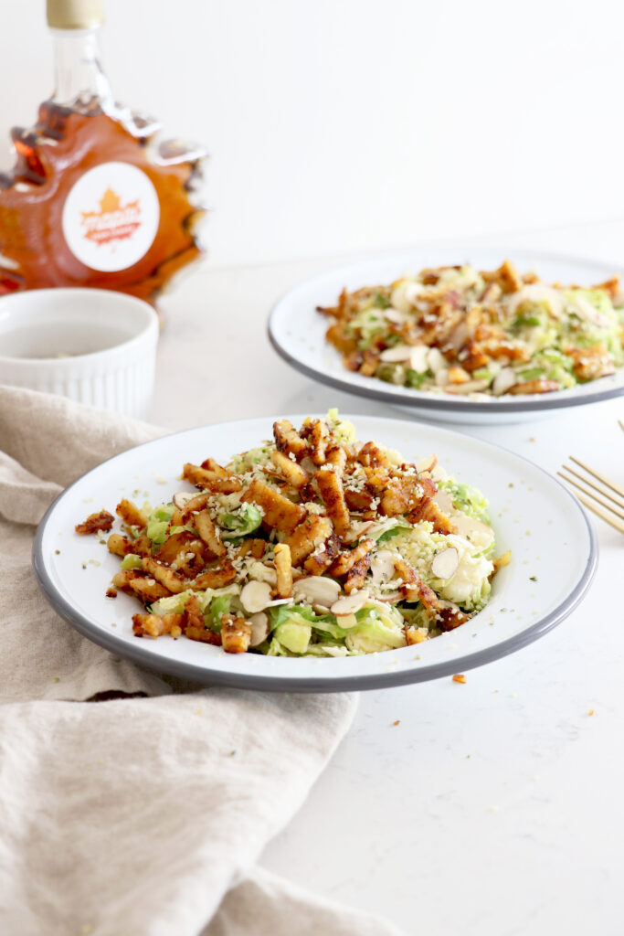 Two white plates of tempeh bacon salad with maple syrup in the back.