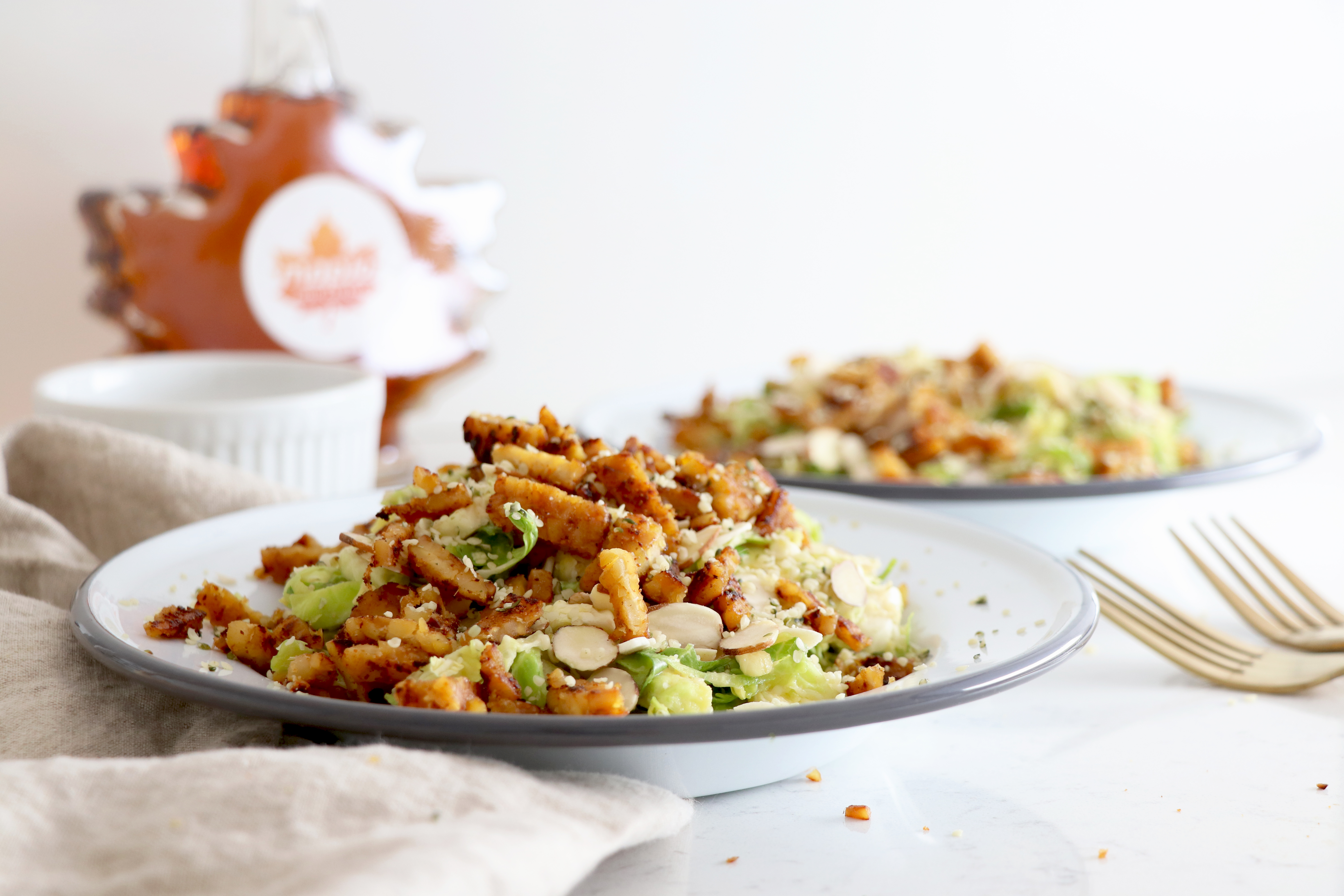 Plates with brussel sprout tempeh bacon salad and syrup bottle in the back.