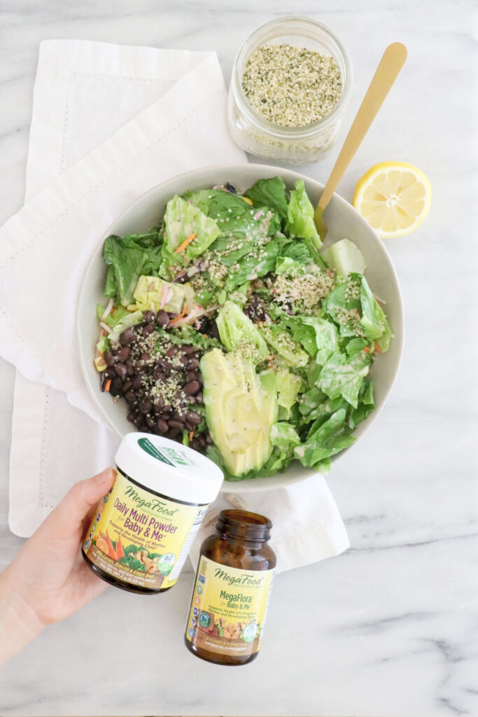 White salad bowl in the middle of photo on white napkin. Gold fork inside bowl. Megafood supplements on the bottom on photo. Lemon slice and hempseed at the top of photo.