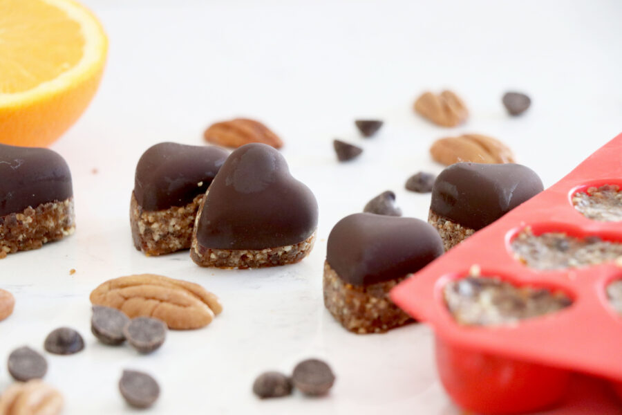 Heart mold on the right with chocolate hearts on the left surrounded by chocolate chips and pecans. Sliced orange in top left corner.
