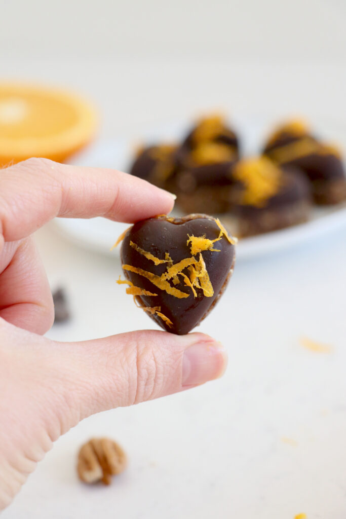 Sliced orange in the background. White plate with chocolate hearts also in background. Front of photo is a hand holding one of the chocolate hearts.