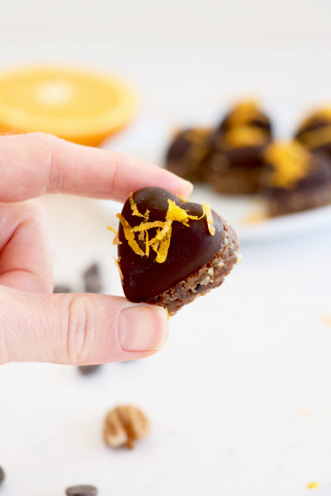 White plate with orange chocolate hearts in background along with sliced orange. Front of photo is a hand holding one chocolate heart.