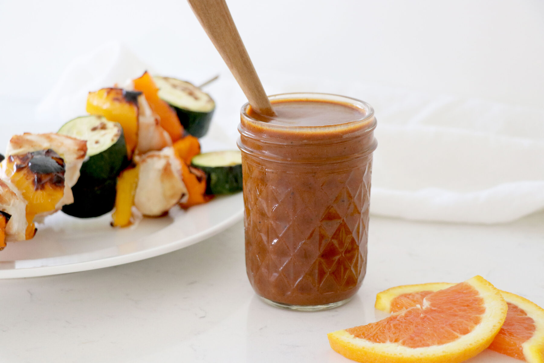 Glass container with bbq sauce and spoon coming out of jar. Grilled food on left side and sliced oranges on bottom right.