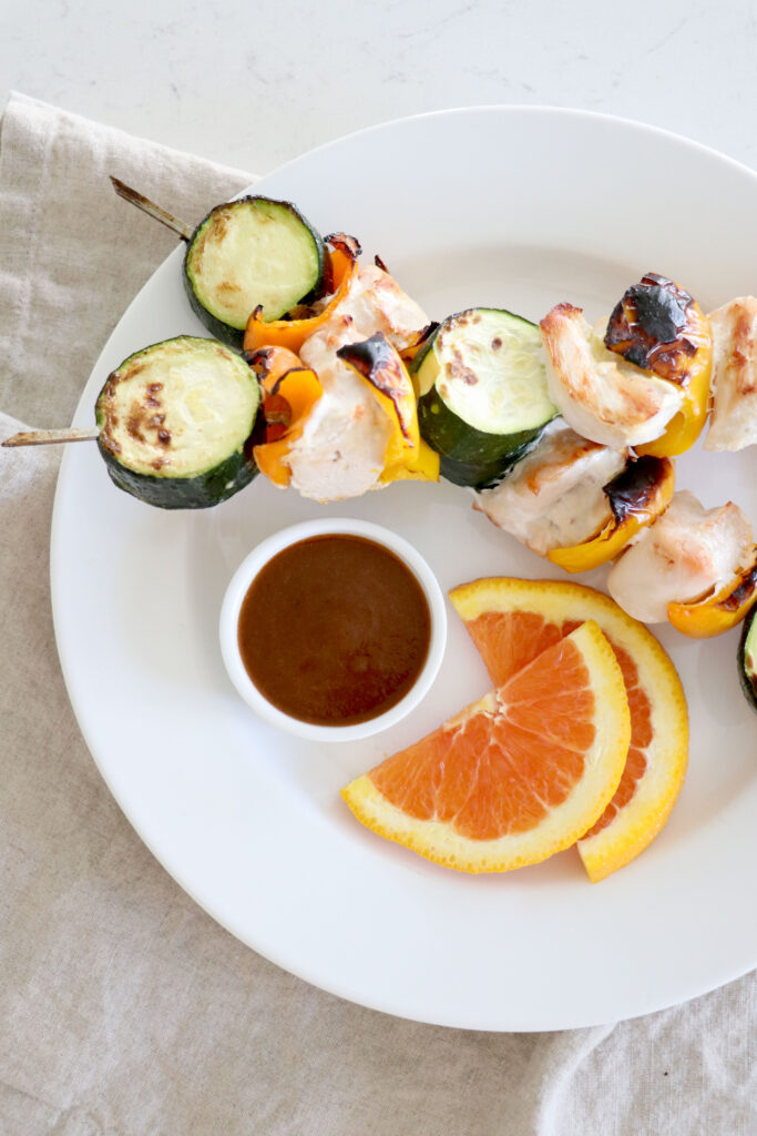 white plate with grilled skewers and a cup of bbq sauce. Orange slices on the side of plate.