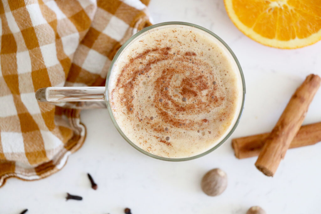 Birds eye view of pumpkin spice latte in glass mug with orange slice, cinnamon sticks and napkin surrounding it.