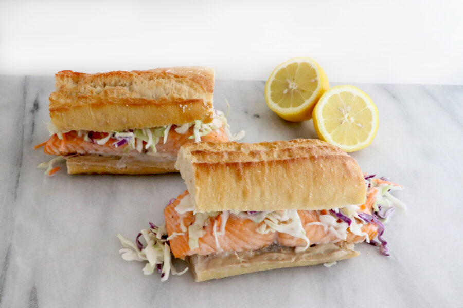 two salmon sandwiches in photo topped with a coconut citrus slaw. Sliced lemon on the right side above sandwich.