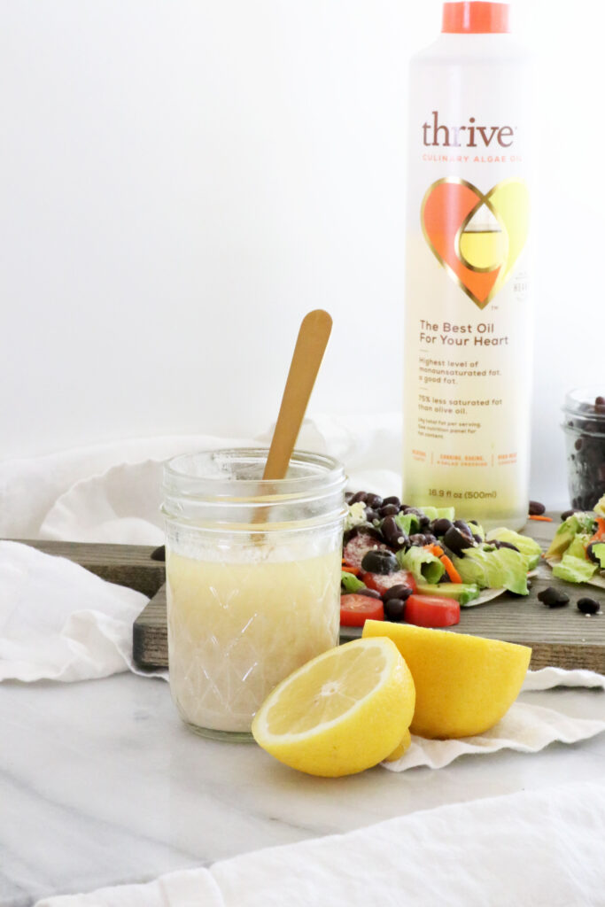 Lemon Tahini Dressing in center of photo with spoon in glass container. Half of lemon next to dressing. Thrive algae oil in picture and salad taco on the side.
