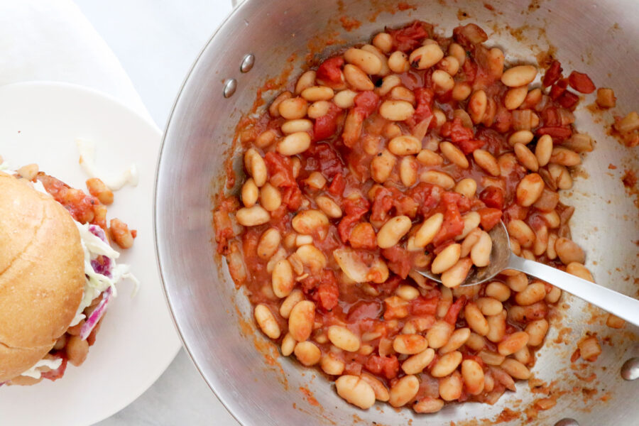 Saucepan on the center and right side of photo filled with bbq bean sloppy Joe mixture. Sandwich on a white plate on the left side of picture.